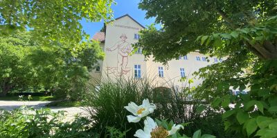Ein helles Schulgebäude mit dem Schriftzug „Landwirtschaftsschule“ ist zwischen dichten, grünen Bäumen und Sträuchern zu sehen. 