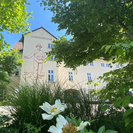 Ein helles Schulgebäude mit dem Schriftzug „Landwirtschaftsschule“ ist zwischen dichten, grünen Bäumen und Sträuchern zu sehenen. 